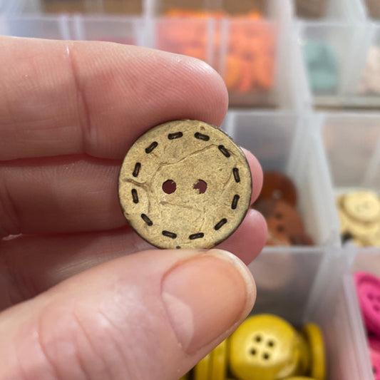 Hand holding a button with a pattern, with a container of buttons in the background.