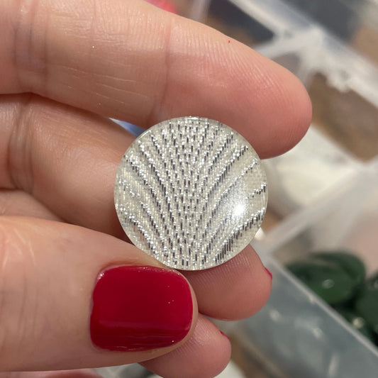 Silver textured earring held by a hand with red nail polish, against a background of jewelry in a box.
