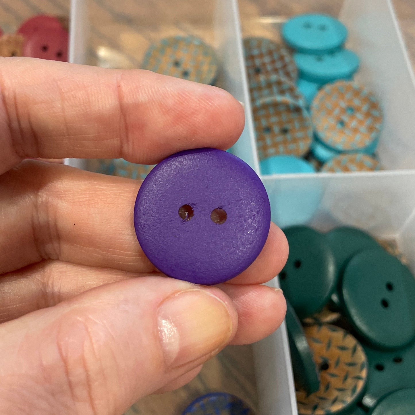 Hand holding a purple button with a container of assorted buttons in the background