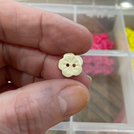 Hand holding a small yellow flower-shaped object with a clear container of assorted colorful objects in the background.