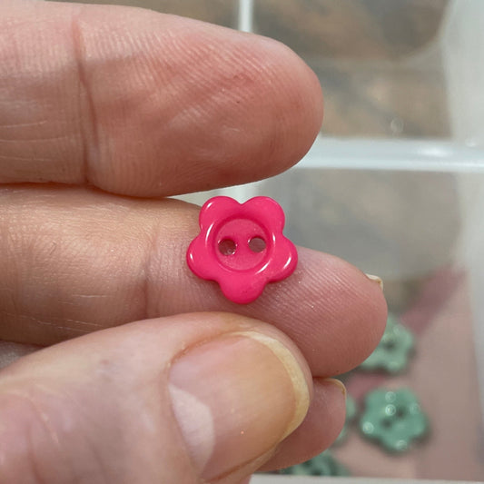 Hand holding a pink flower-shaped object with a container of buttons in the background