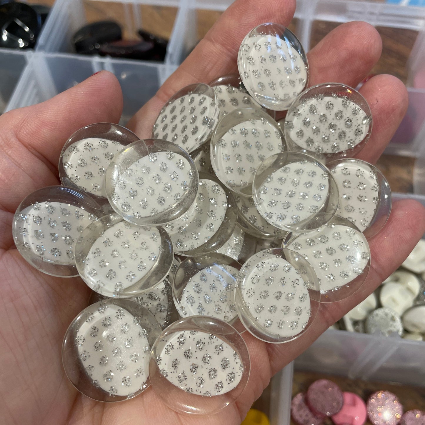 Hand holding clear round objects with embedded materials, likely for crafting or jewelry making, with a blurred background of similar items.