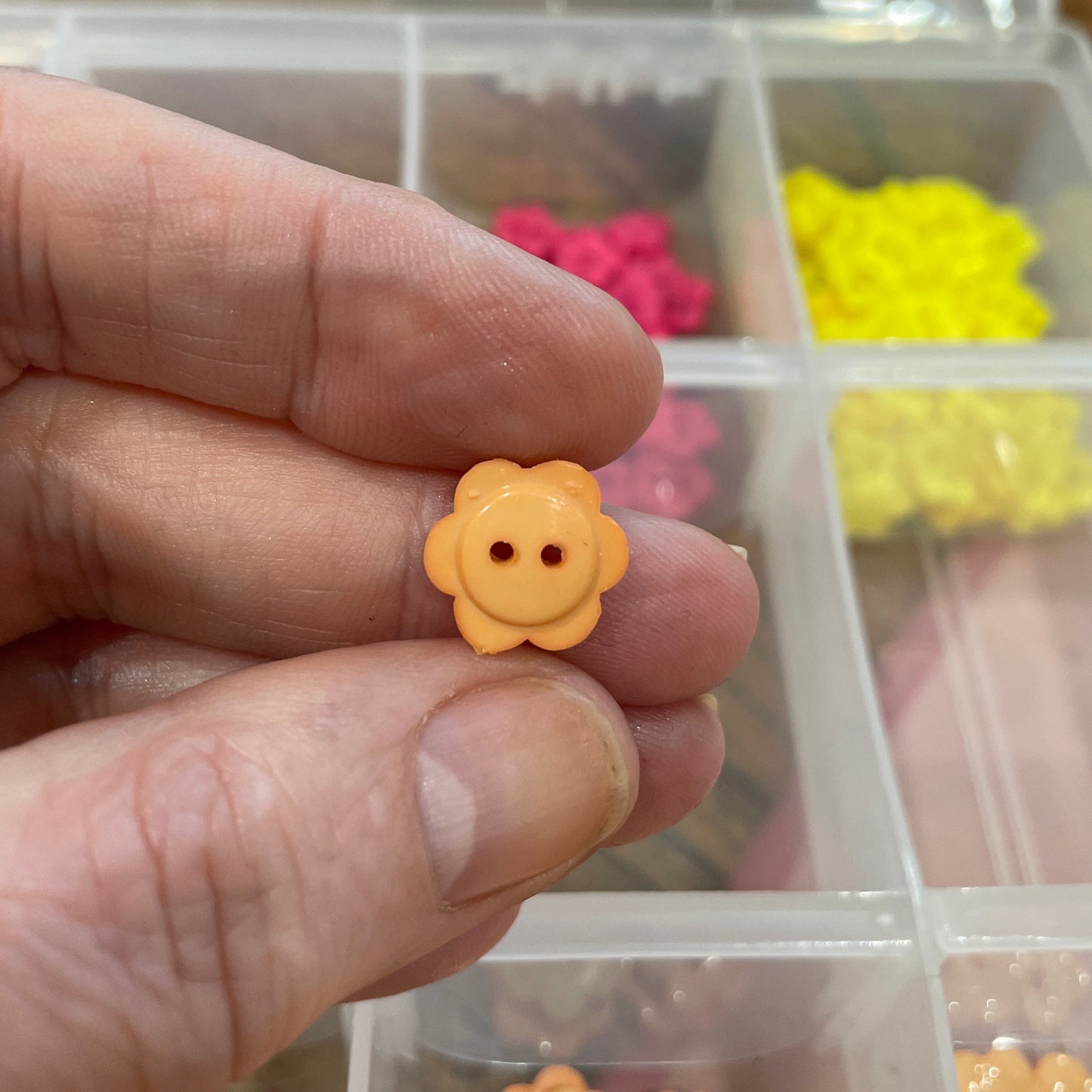 Hand holding a small yellow flower-shaped button with a blurred container of buttons in the background.