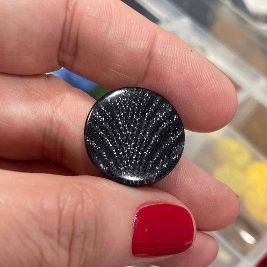 Hand holding a black textured button with a blurred background of a jewelry box.