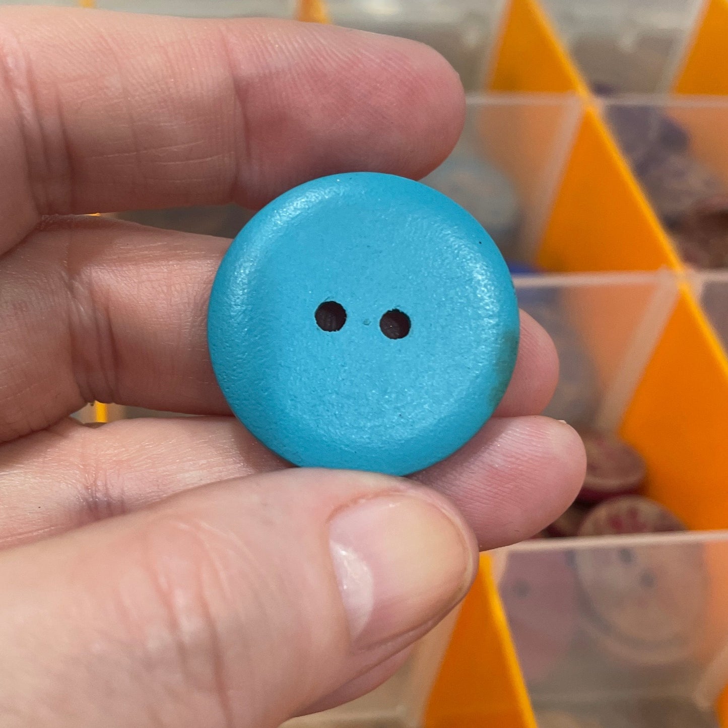 Hand holding a blue button with a clear container of buttons in the background
