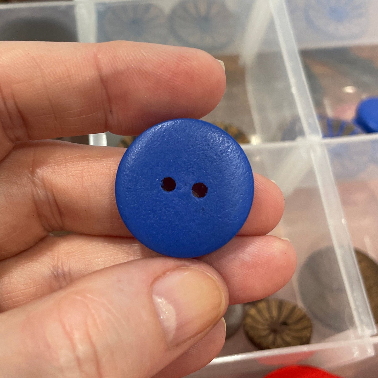 Hand holding a blue button with a clear container of buttons in the background