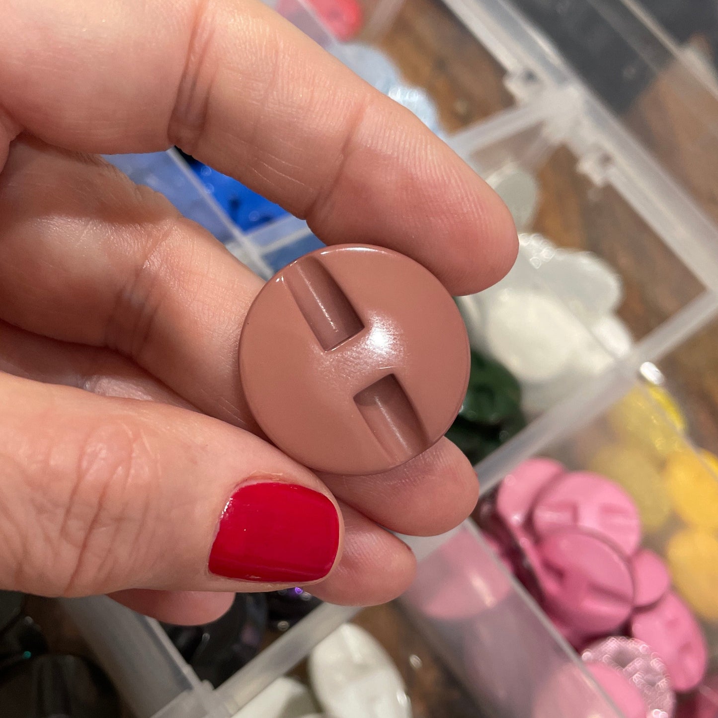 Hand holding a round beige button with a clear container of various buttons in the background.