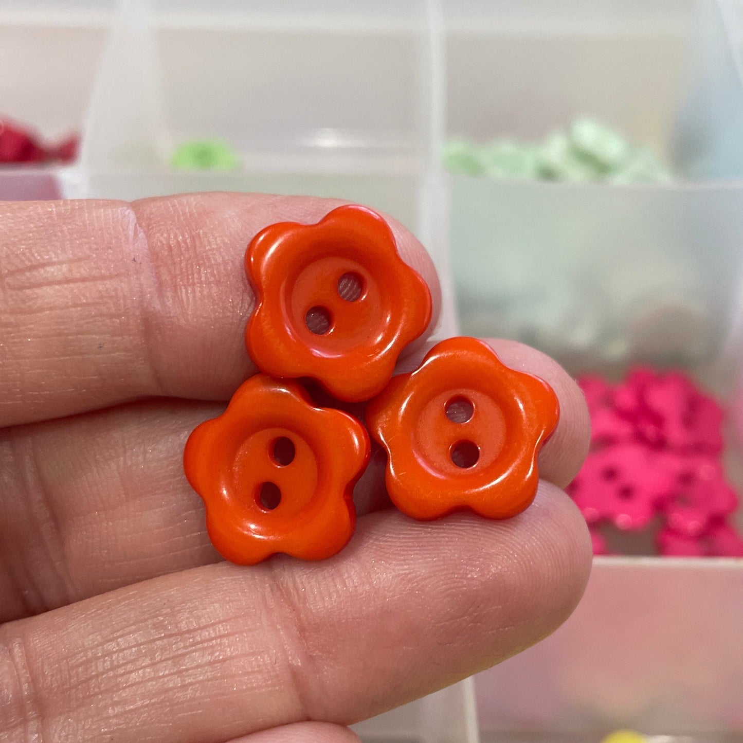 Hand holding two orange flower-shaped buttons with a background of colorful buttons in a box.