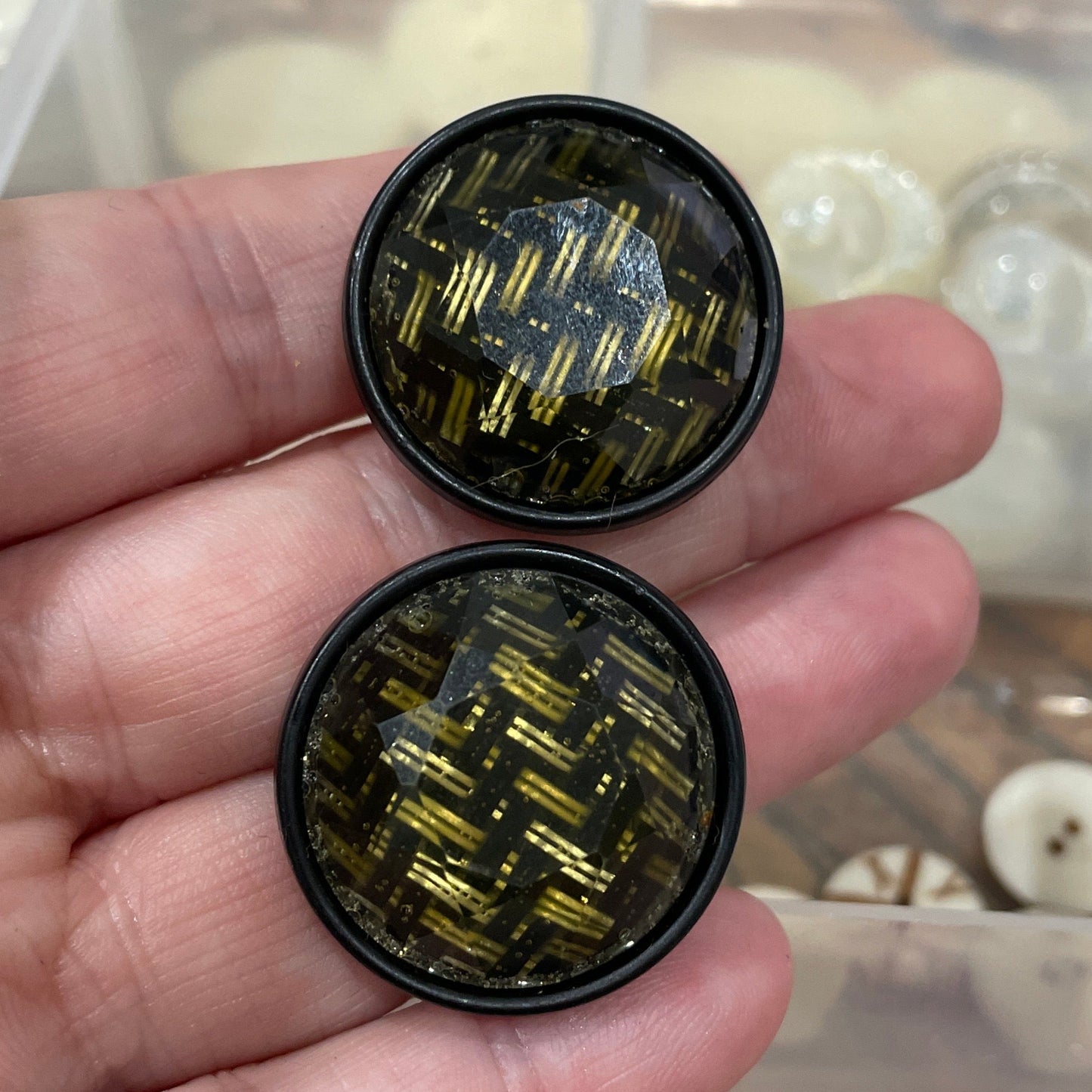 Two black and gold circular objects held in a hand with a blurred background of beads.