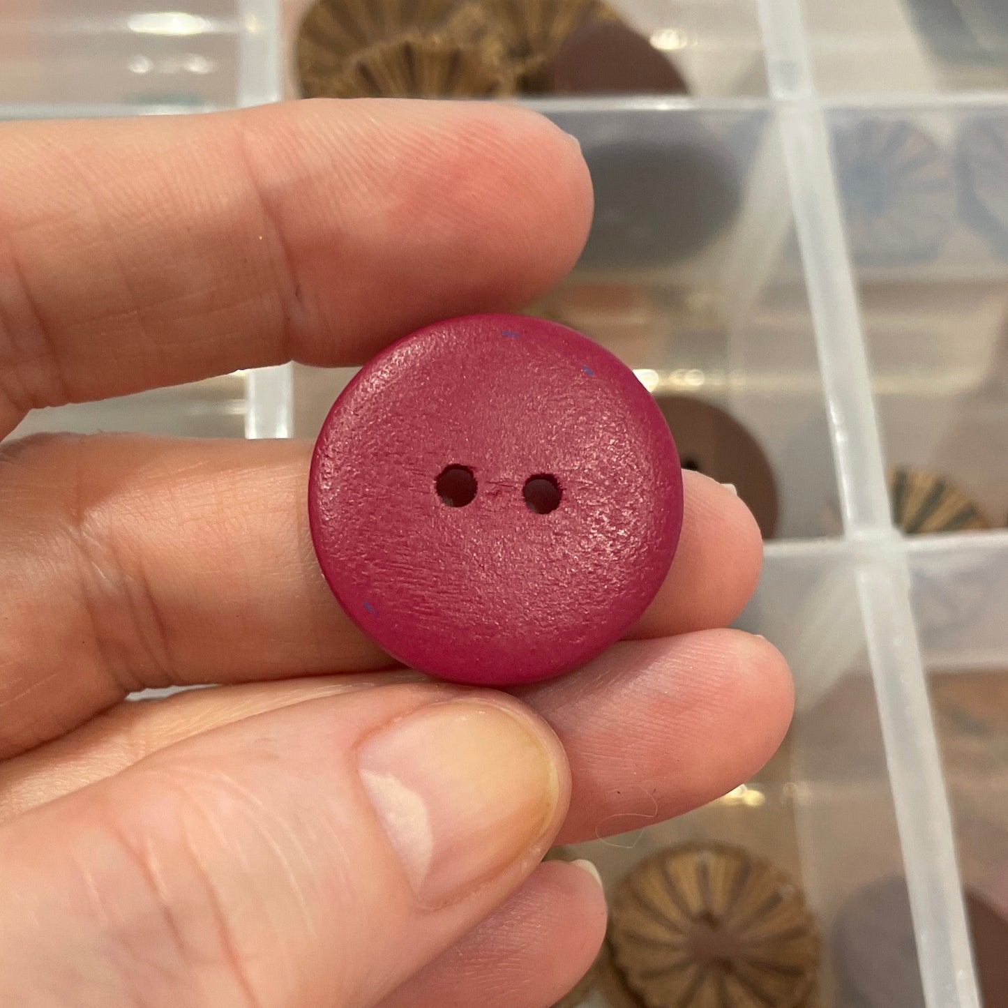 Hand holding a pink button with a background of assorted buttons in a clear container.