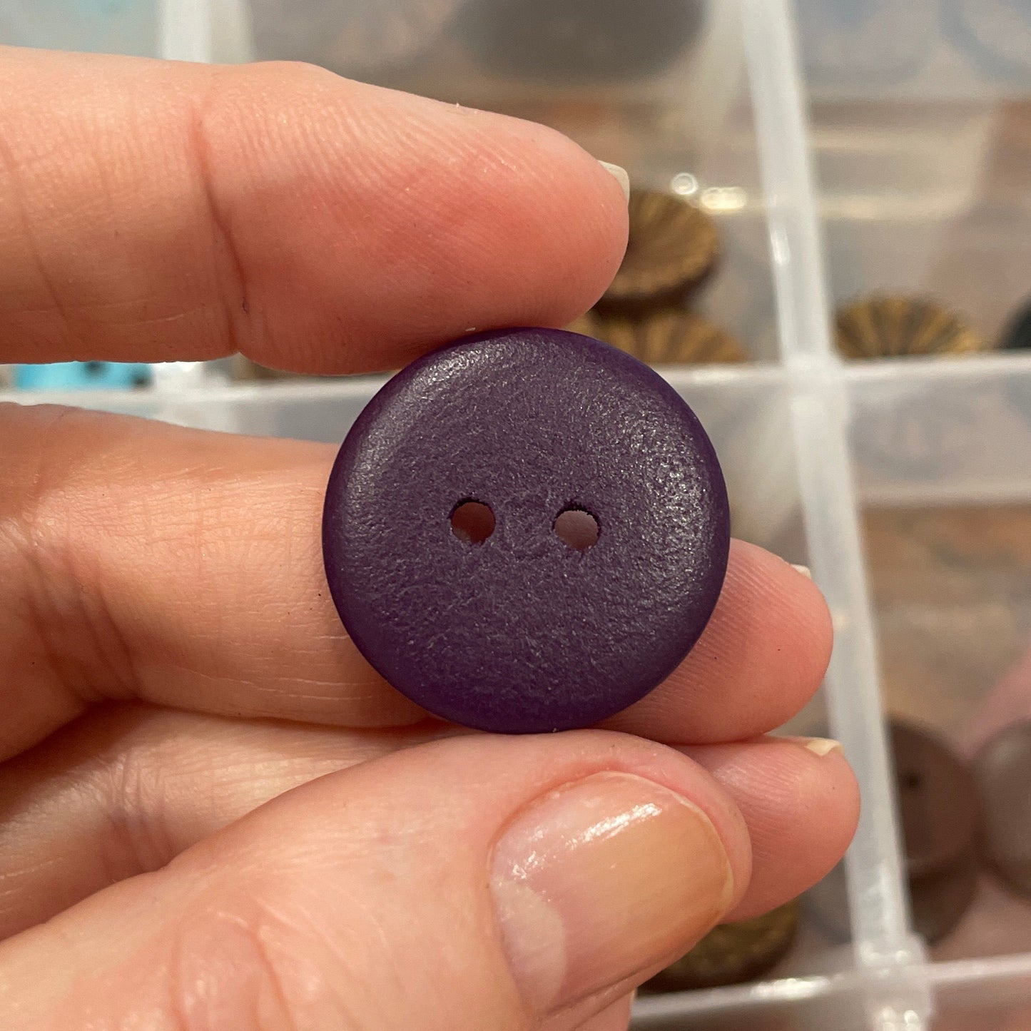 Hand holding a purple button with a clear container of assorted buttons in the background