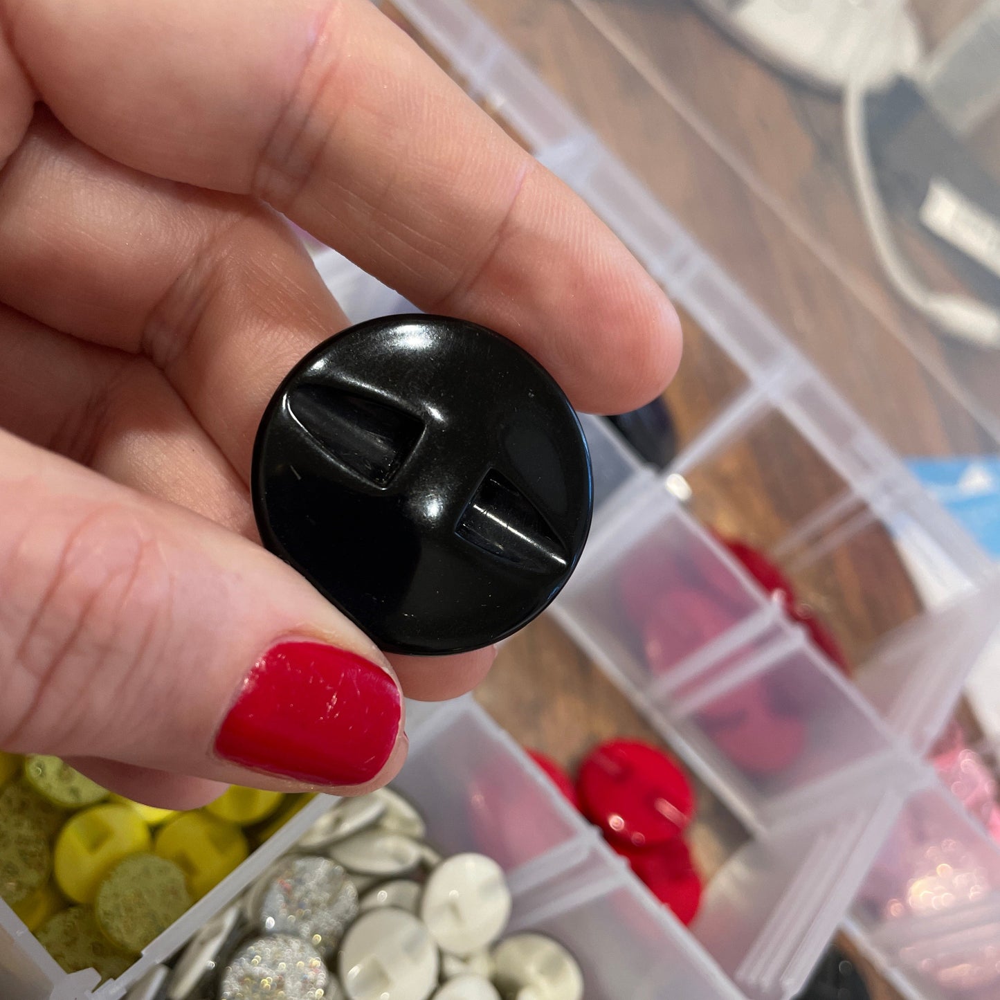 Black button held by a hand with red nail polish, in front of a container of assorted buttons.