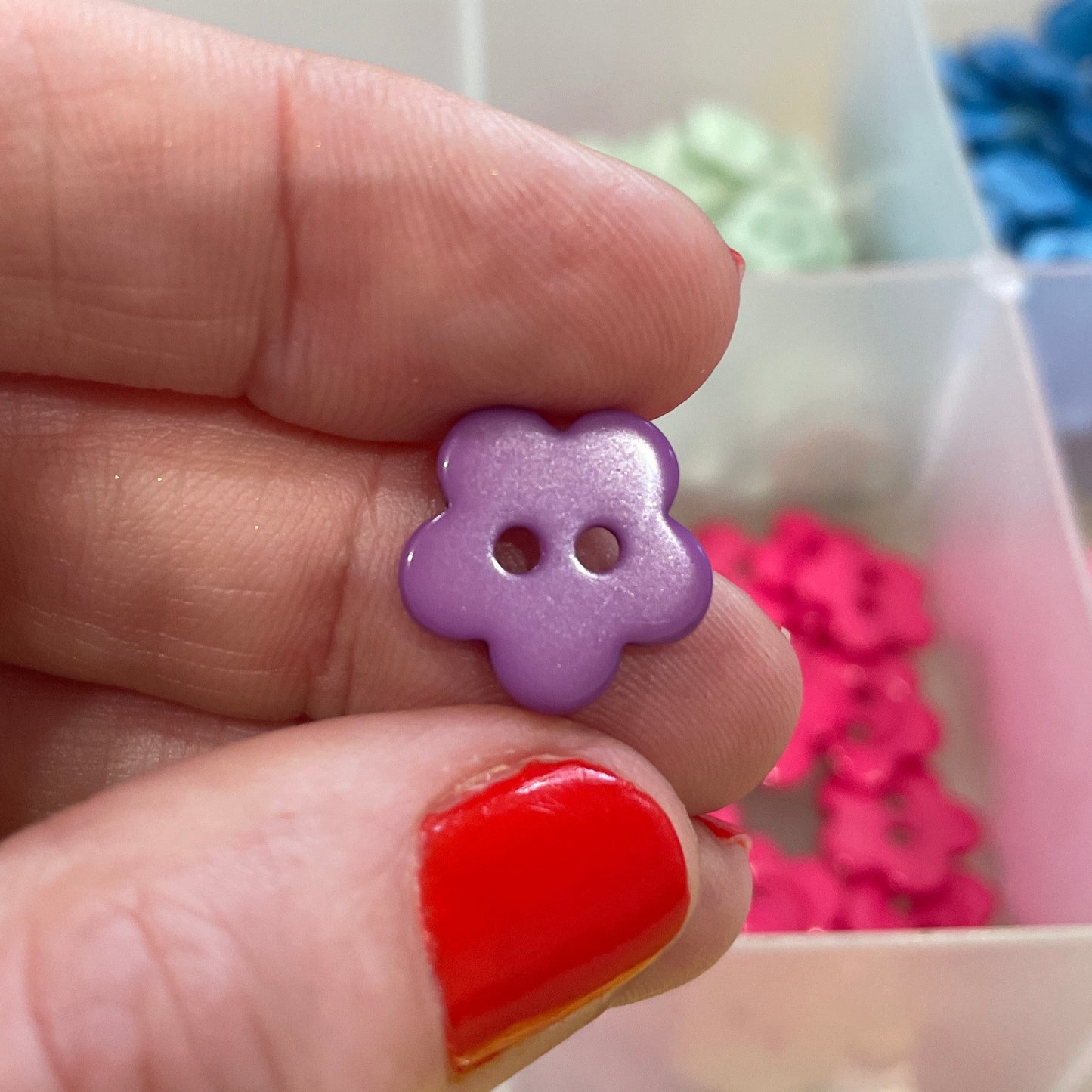 Hand holding a purple flower-shaped button with a background of colorful buttons in a box.