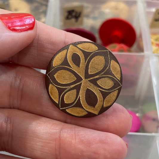 Hand holding a decorative wooden button with a detailed pattern, against a background of various buttons in a container.
