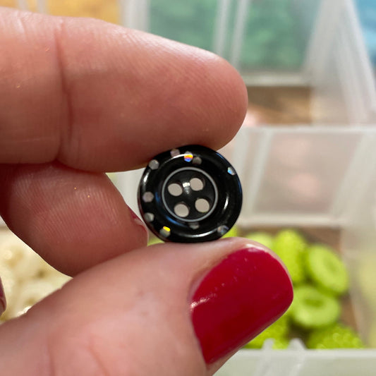 Black button held by a hand with red nail polish, against a background of various buttons in a container.