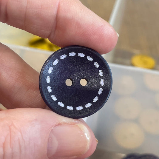 Hand holding a black button with white dots in front of a container of various buttons.