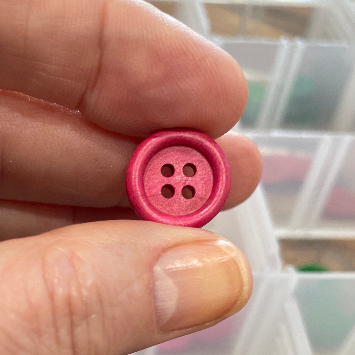 Hand holding a pink button with a container of buttons in the background