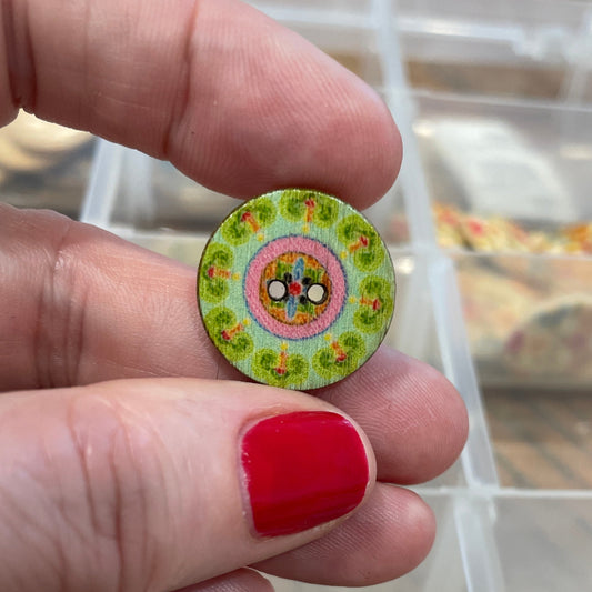 Hand holding a small round green button with floral design, against a blurred background of similar buttons.