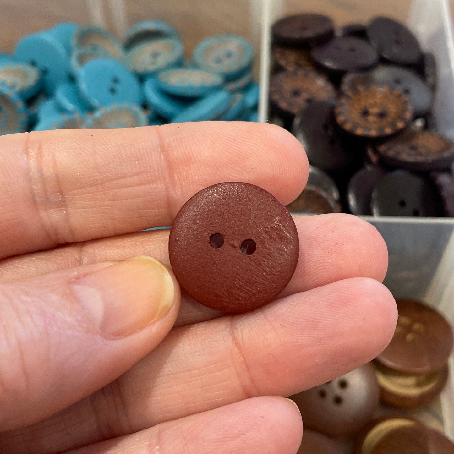 Hand holding a brown button with a clear container of buttons in the background