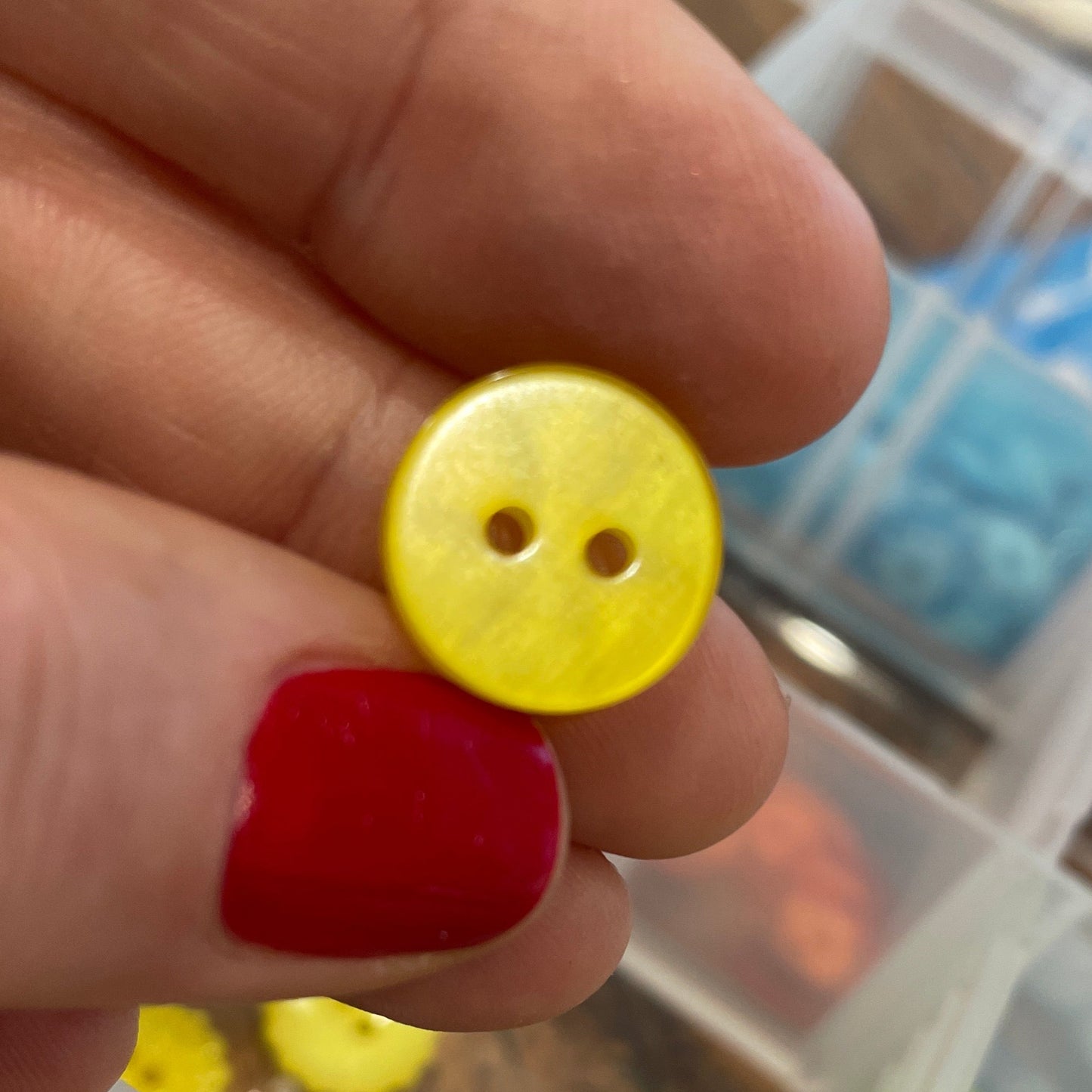 Yellow button held by a hand with red nail polish, against a background of colorful buttons in a case.