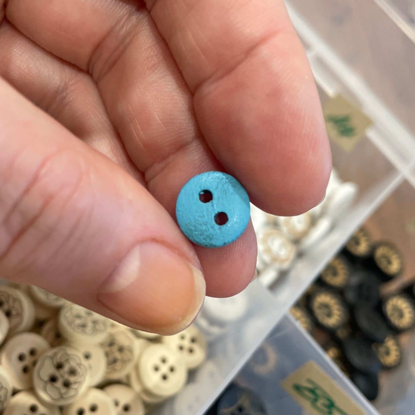 Hand holding a small blue button with a container of various buttons in the background
