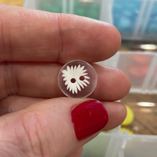 Hand holding a small white object with a design, with a container of beads in the background.