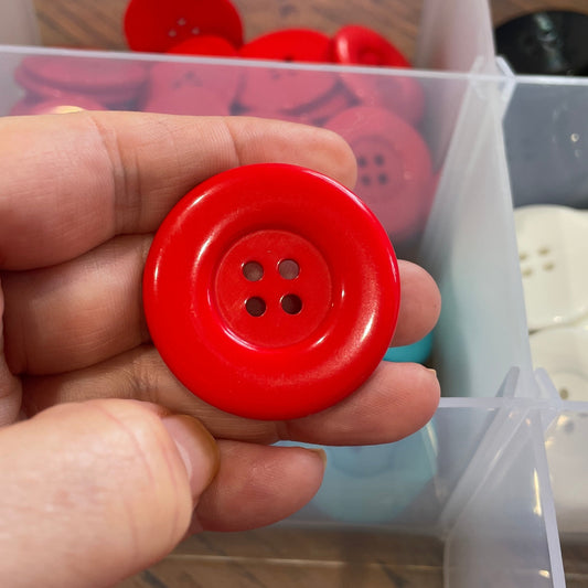 Hand holding a red button with a clear container of buttons in the background