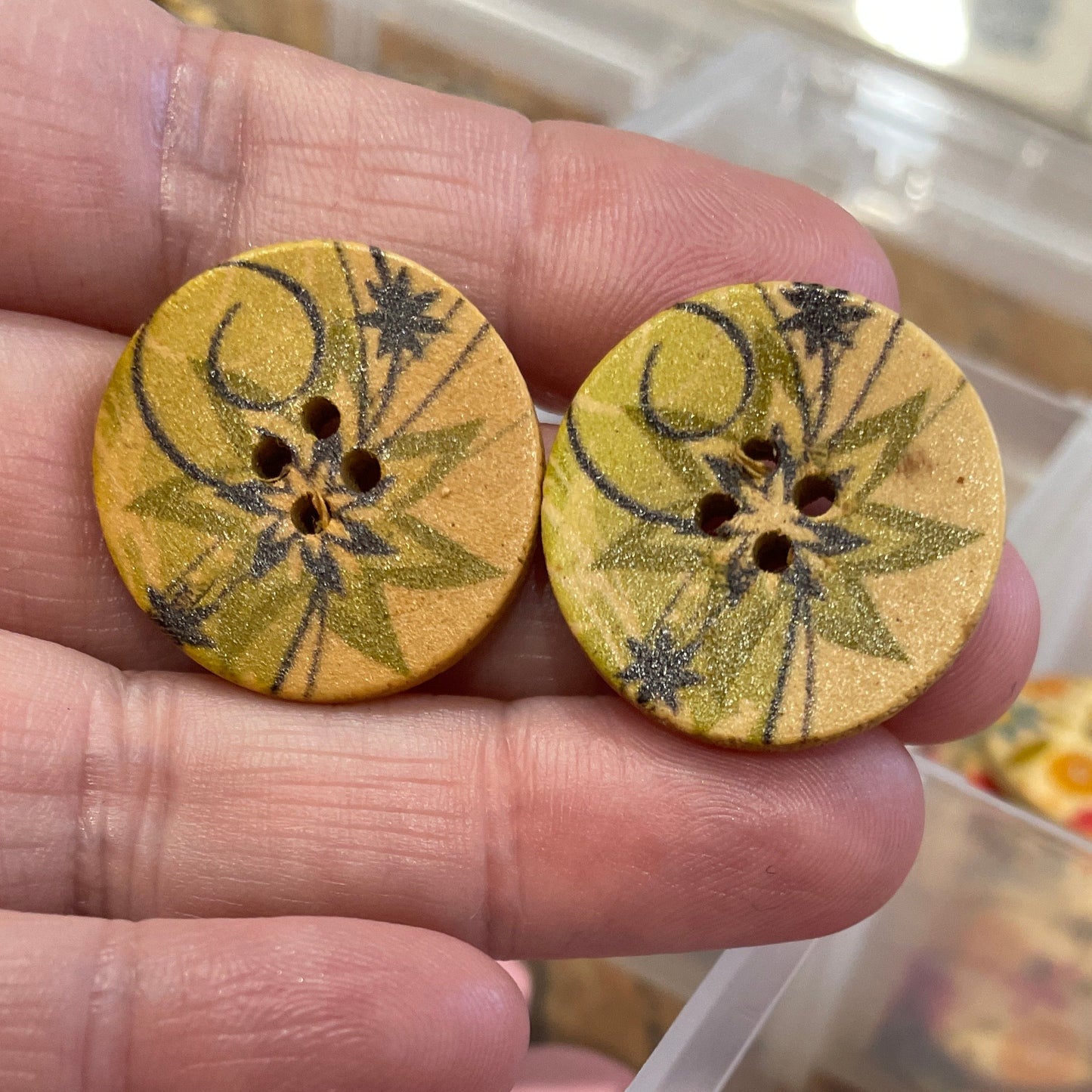 Two yellow floral buttons held between fingers with a container of assorted buttons in the background.