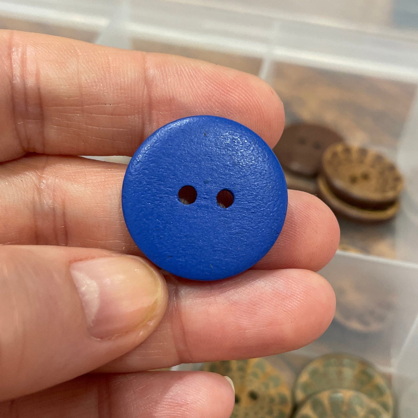 Hand holding a blue button with a blurred background of assorted buttons.