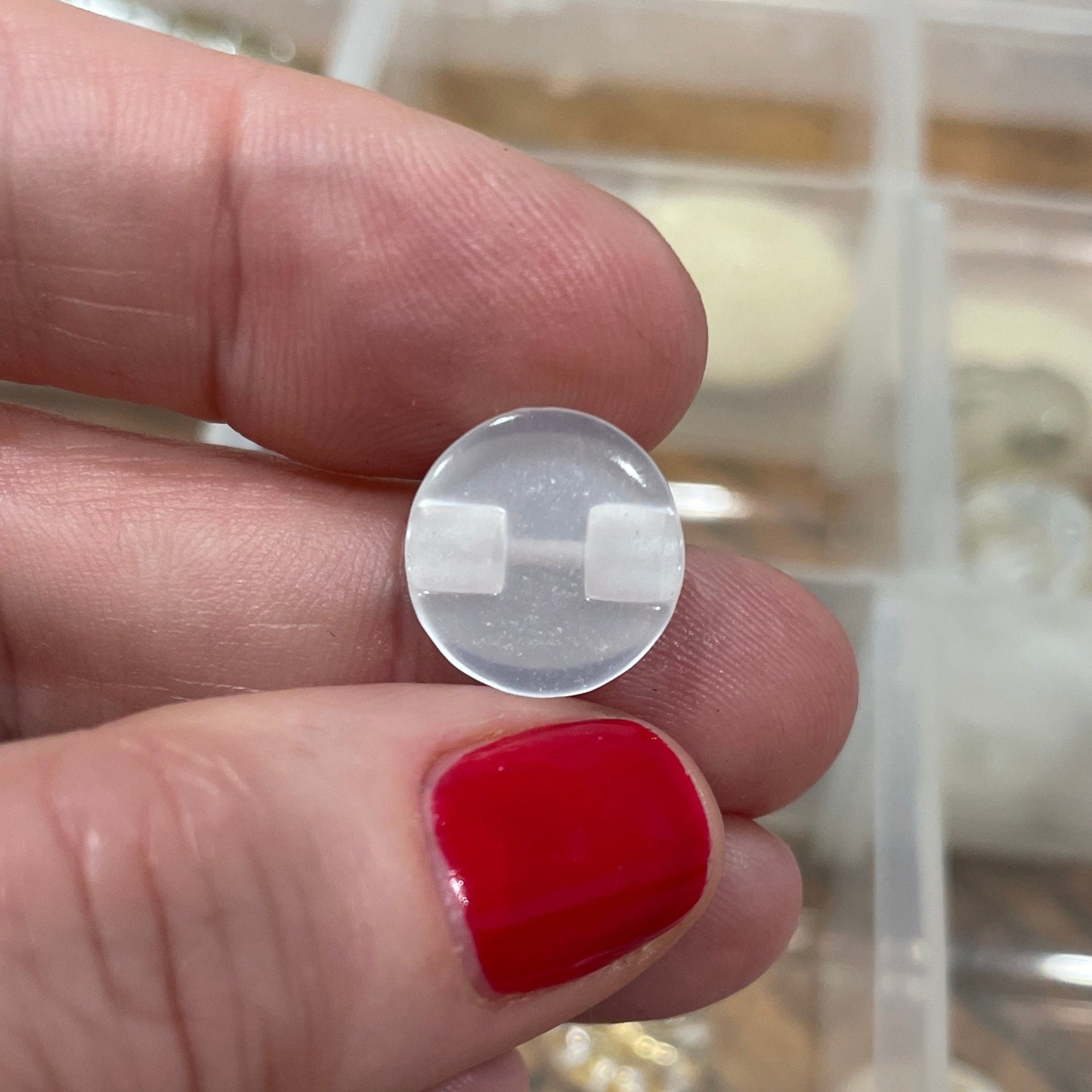 Hand holding a clear circular object with a grid pattern, with a container of buttons in the background.