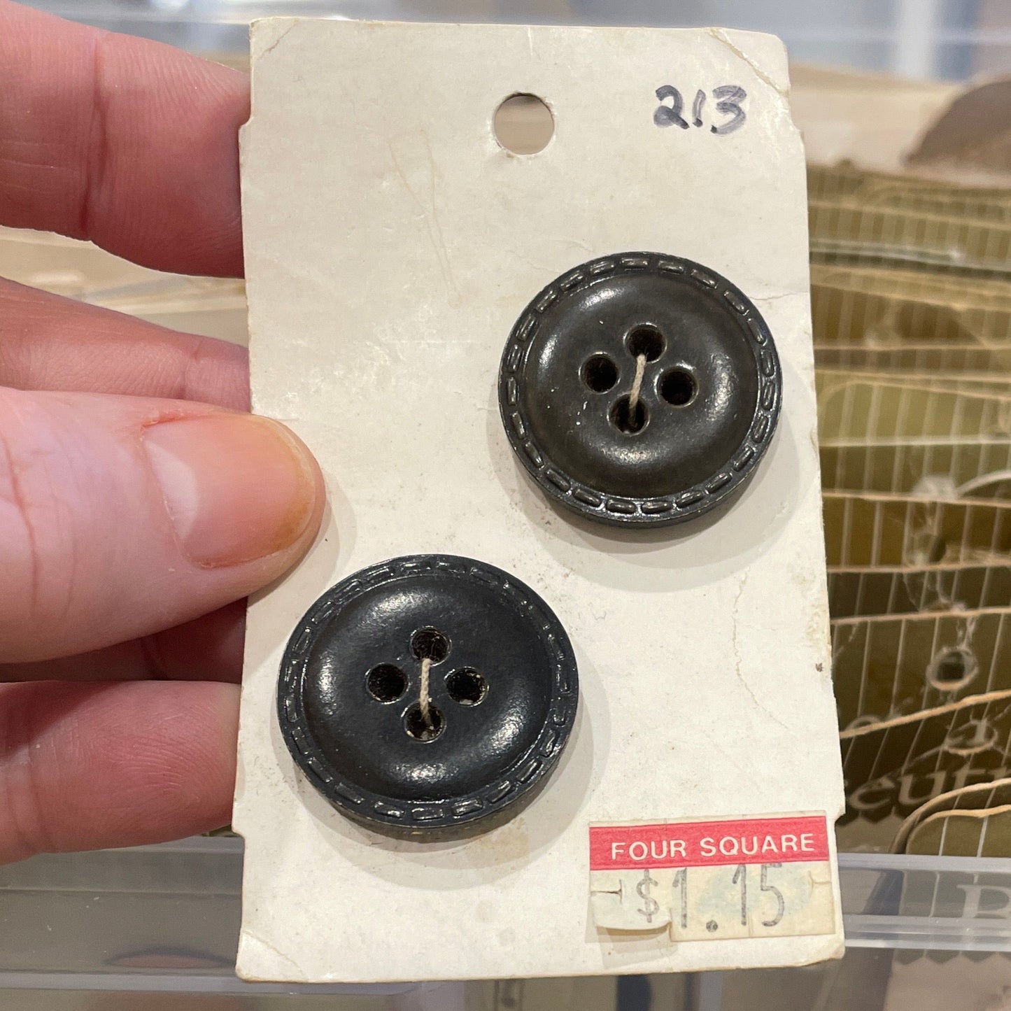 Two black buttons on a card held by a hand with a clear plastic container in the background.