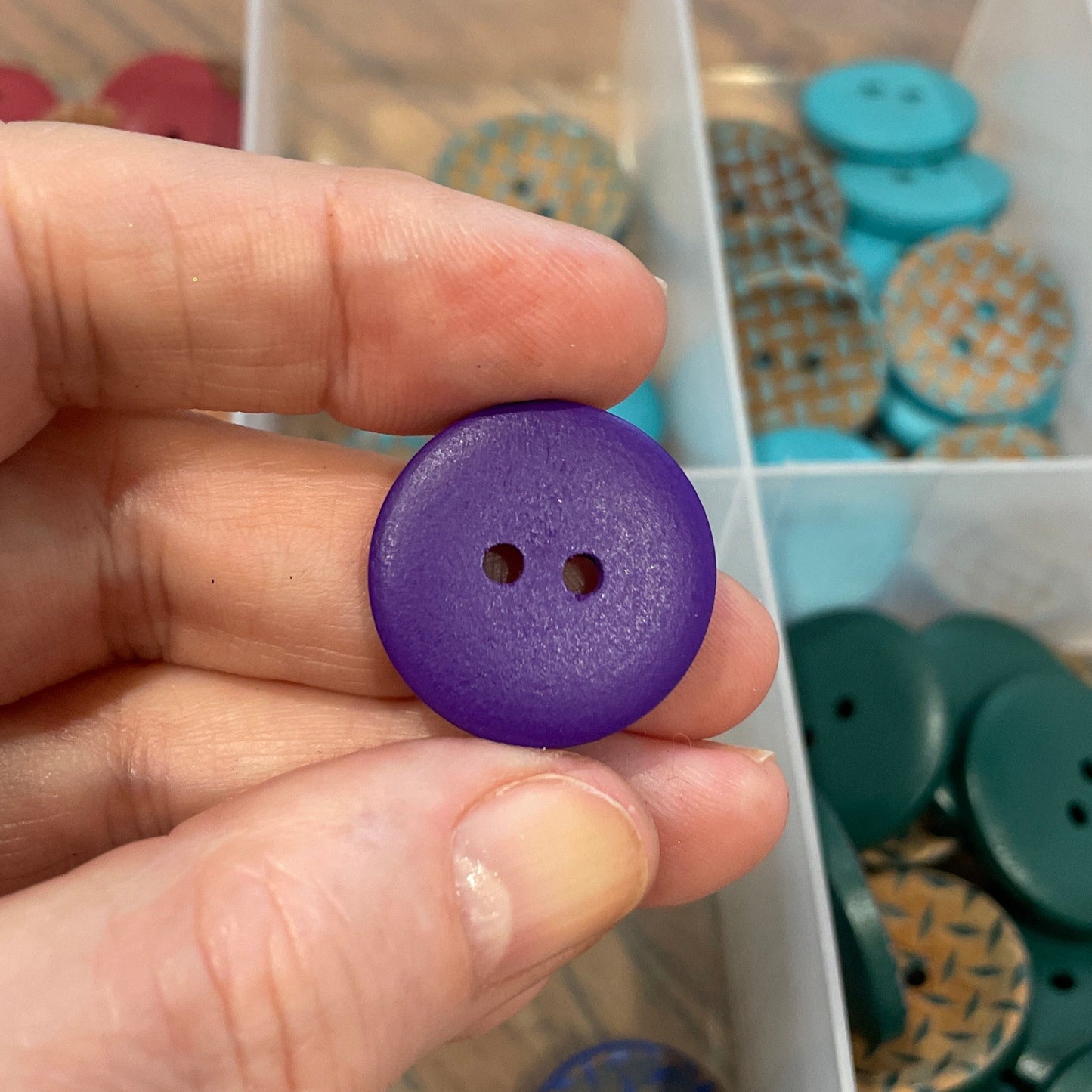 Hand holding a purple button with a clear container of assorted buttons in the background