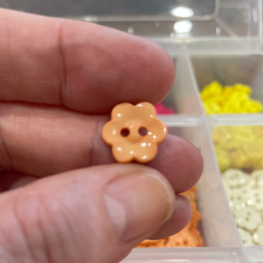 Hand holding a small yellow flower-shaped button with a blurred container of buttons in the background.