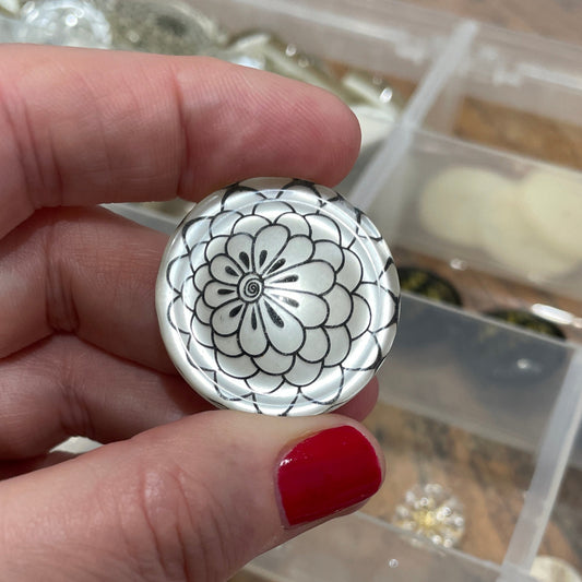 Hand holding a black and white floral design button with a container of beads in the background.
