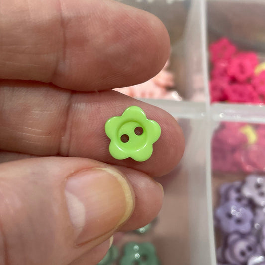 Hand holding a green flower-shaped object with a container of colorful buttons in the background.