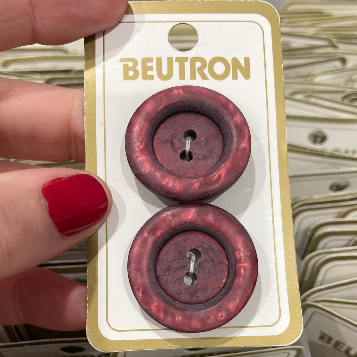 Two maroon buttons on a Beutron card held by a hand with red nail polish.
