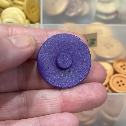 Hand holding a purple button with a container of assorted buttons in the background
