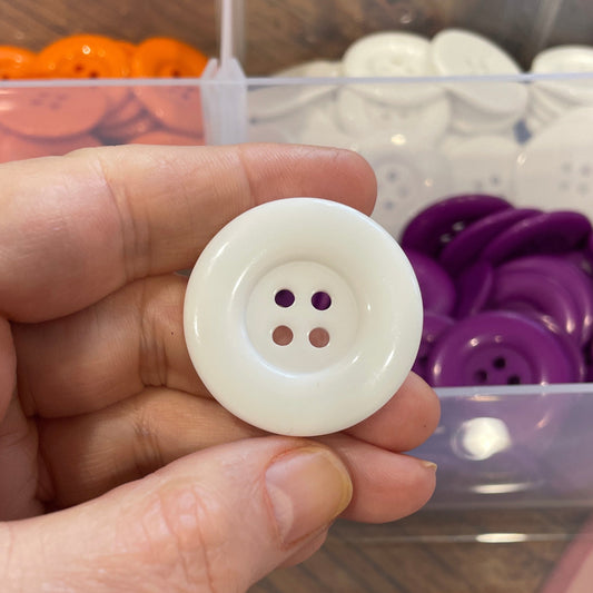Hand holding a white button with a clear container of buttons in the background