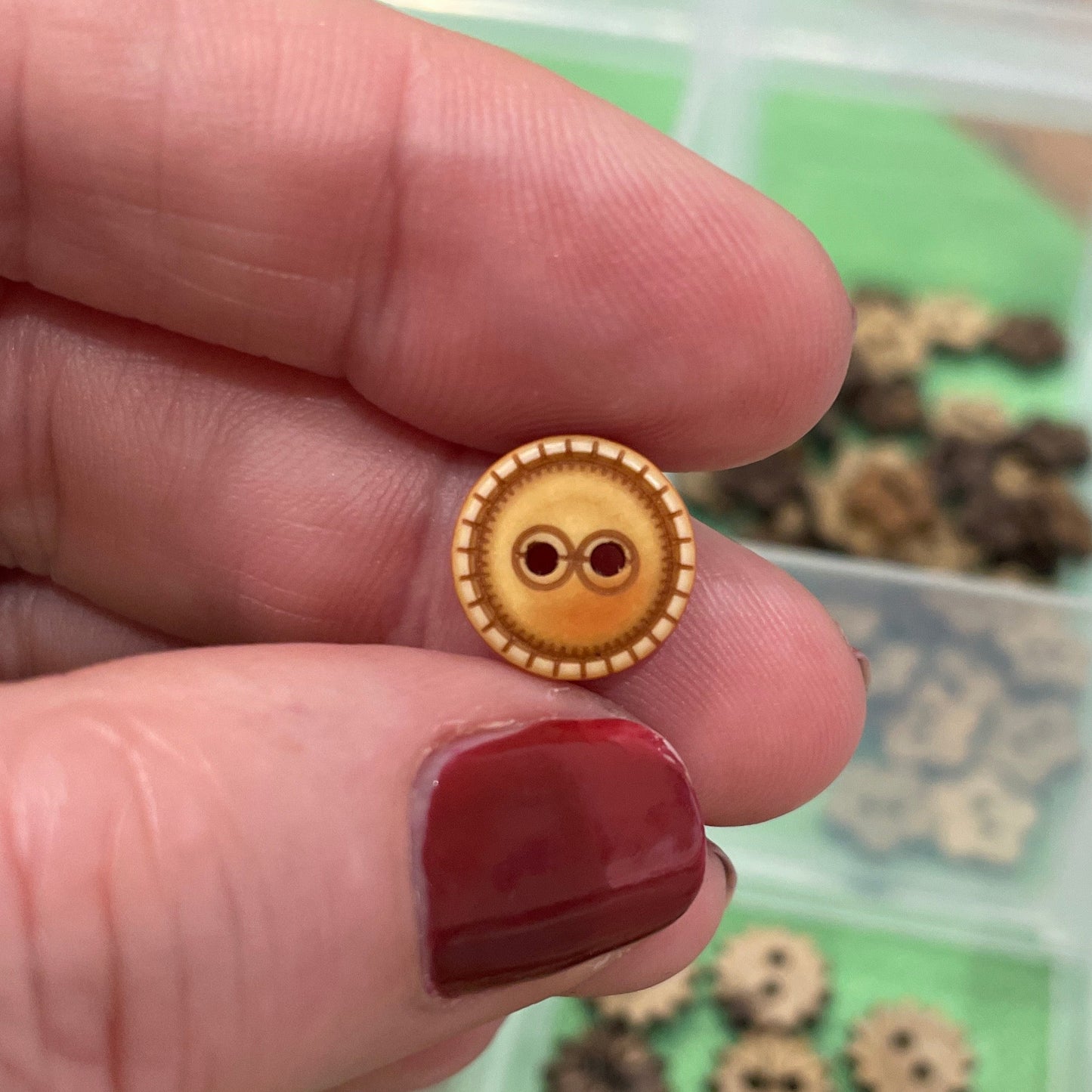 Gold button held by a hand with red nail polish, against a background of more buttons in a container.
