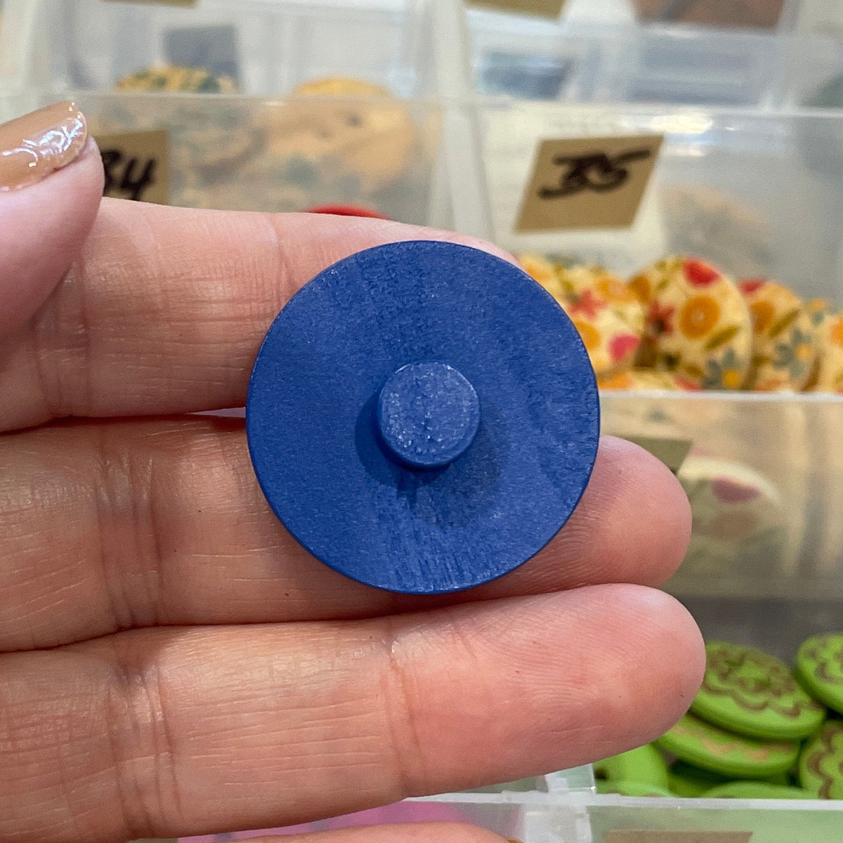 Hand holding a blue button in front of a container filled with various buttons.