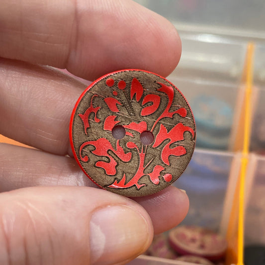Hand holding a decorative button with red and gold patterns in front of a display of buttons.