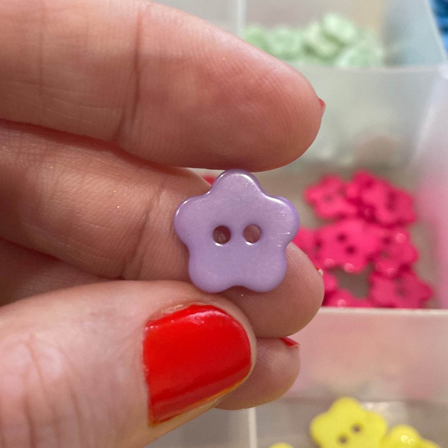 Hand holding a small purple flower-shaped button with a blurred background of colorful buttons.