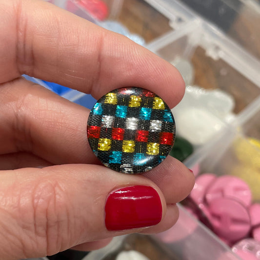 Hand holding a colorful button with a checkered pattern against a background of various buttons in a container.