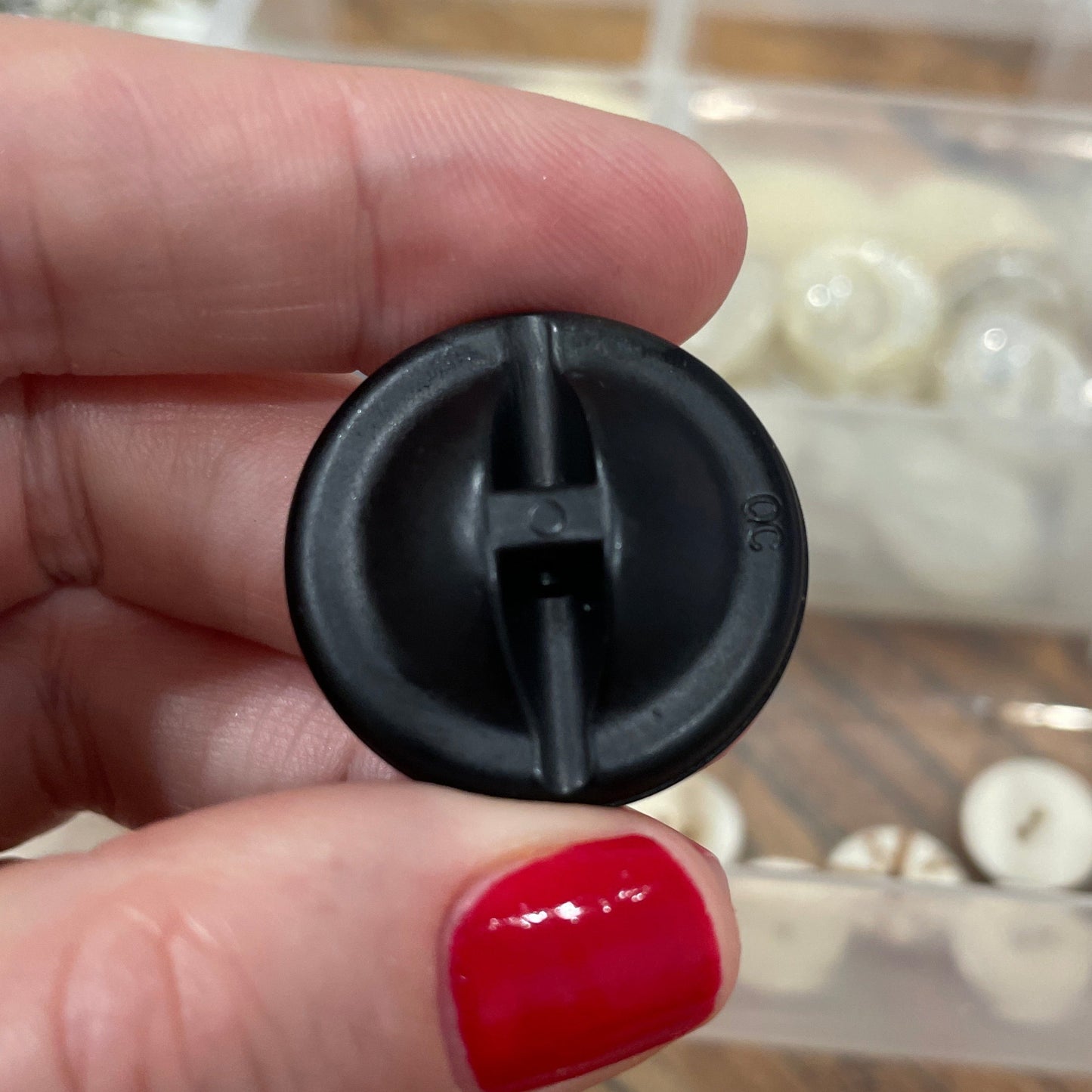 Black plastic component held by a hand with red nail polish, with a clear container of small items in the background.