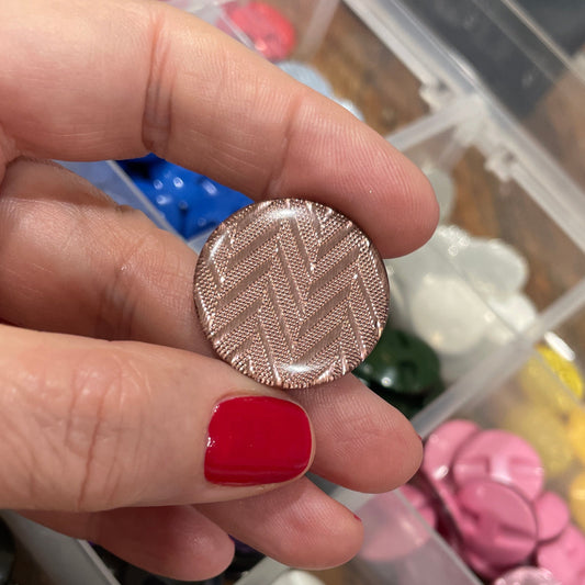Hand holding a textured silver button with a container of various buttons in the background