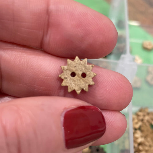 Gold gear-shaped button held between fingers with a container of similar objects in the background.