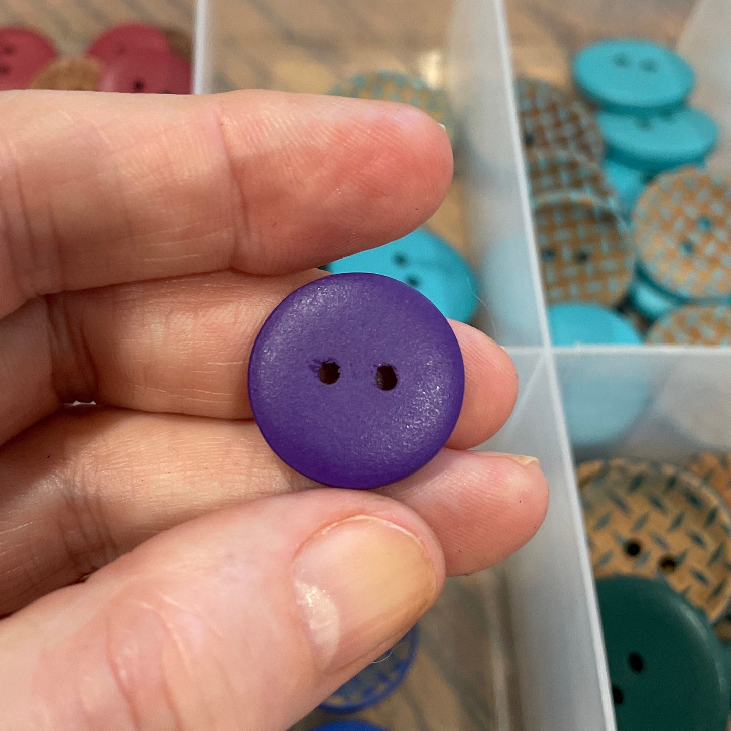 Hand holding a purple button with a clear container of buttons in the background