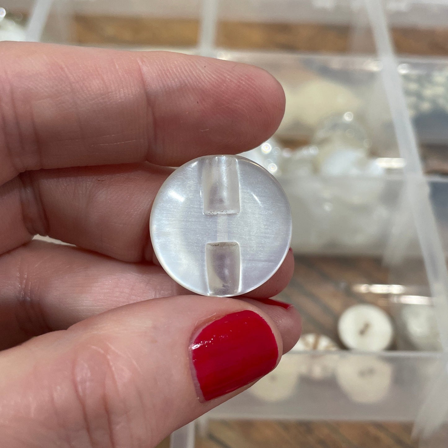 Hand holding a clear plastic component with a container of buttons in the background