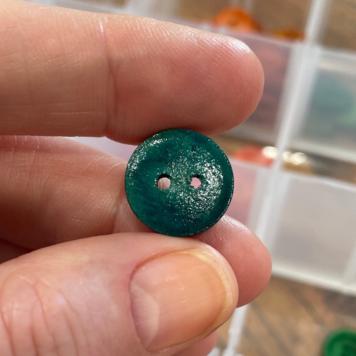 Hand holding a green button with a blurred background of button storage containers.