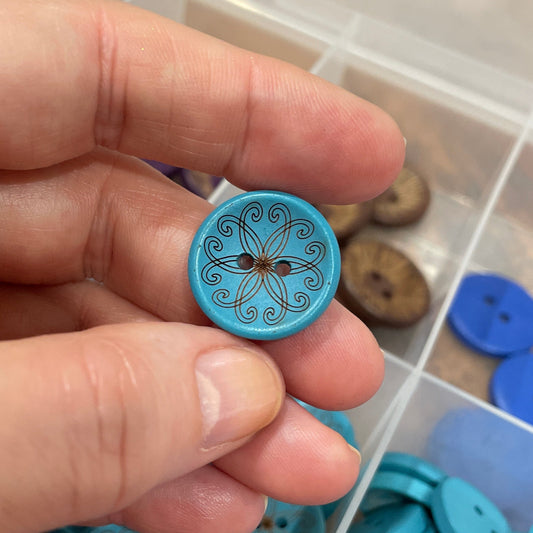 Hand holding a blue button with intricate design, surrounded by more buttons in a box.